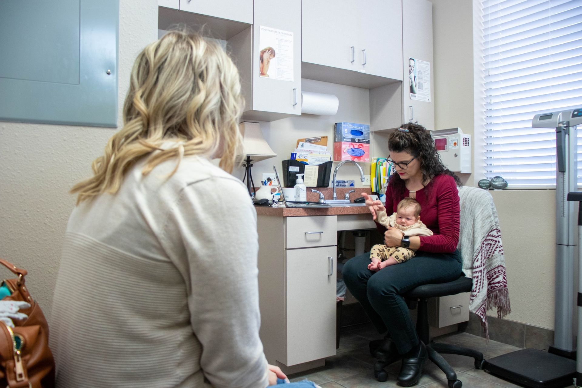 A medical professional sits in an exam room, holding an infant while speaking to a patient seated in the foreground.