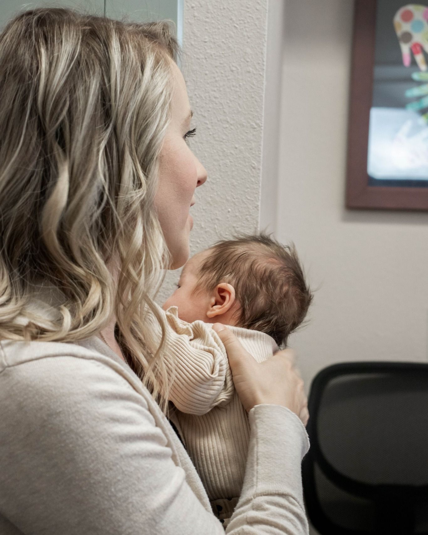 A person with blonde hair holds a newborn infant against their chest in a softly lit, indoor setting.
