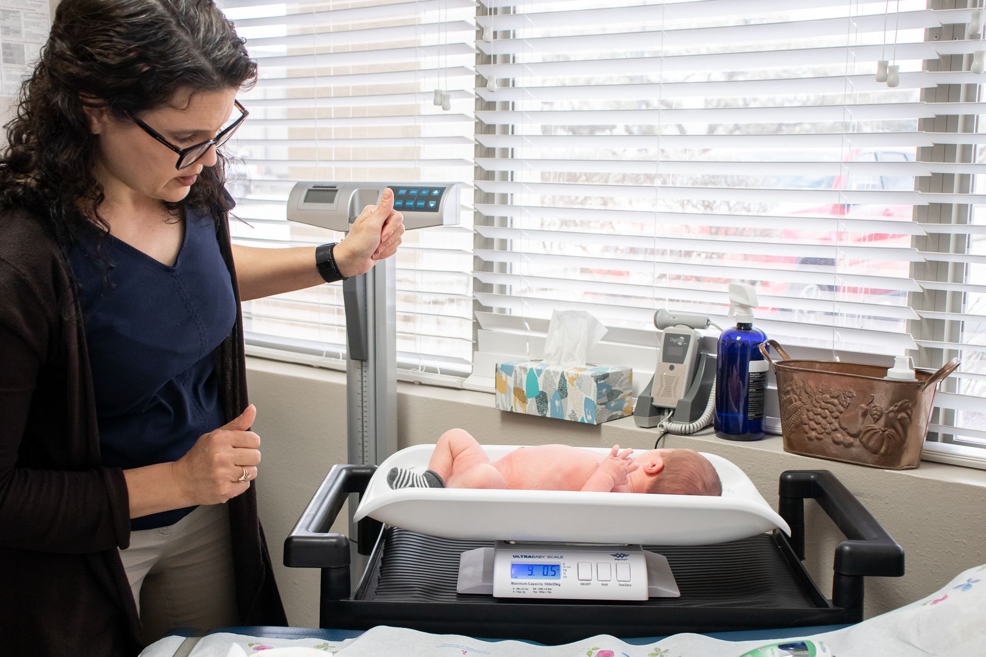 A person stands by a scale, monitoring a newborn baby lying on a medical tray in a clinical room.