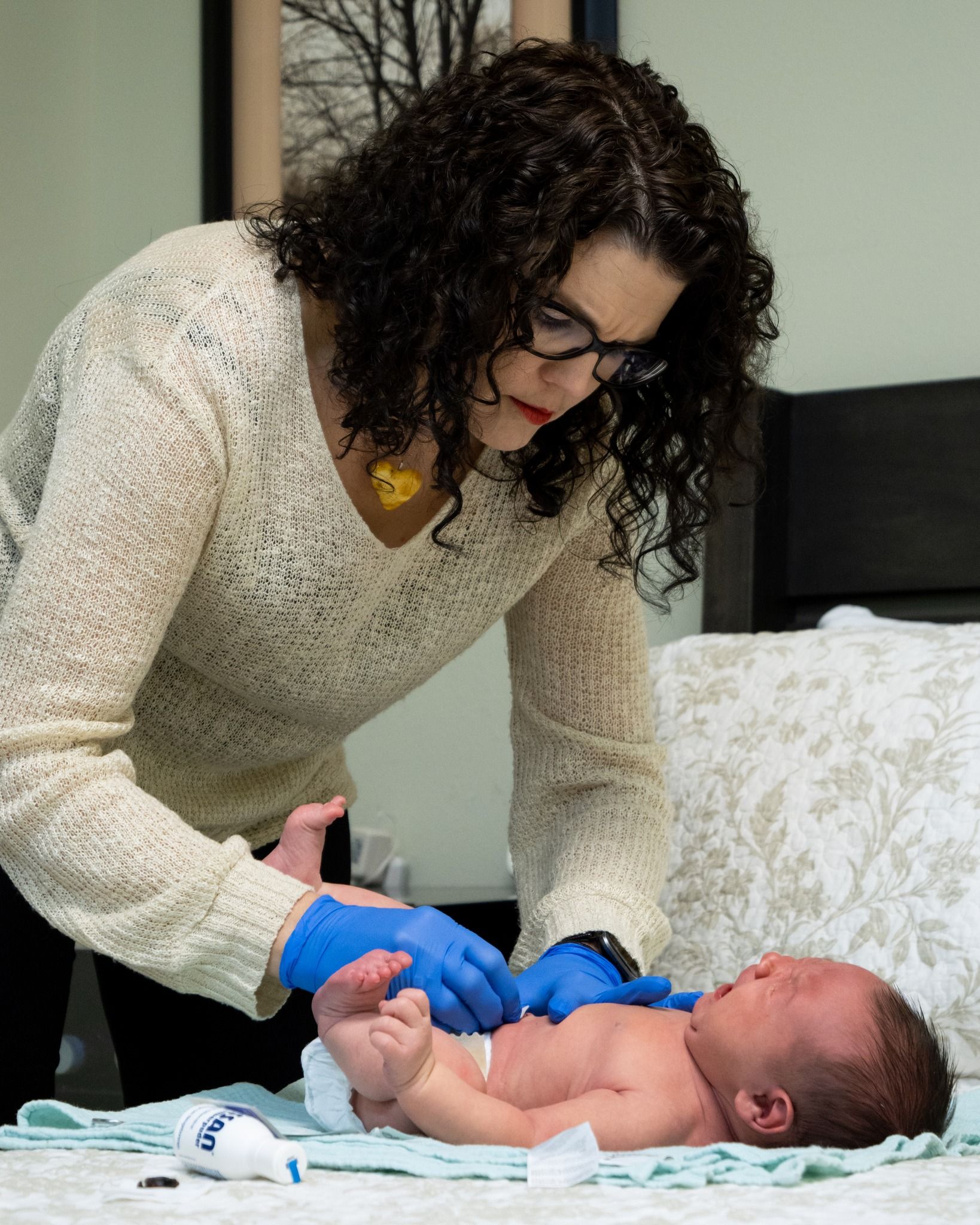 A person in a cream sweater and blue medical gloves tends to a crying newborn on a bed.