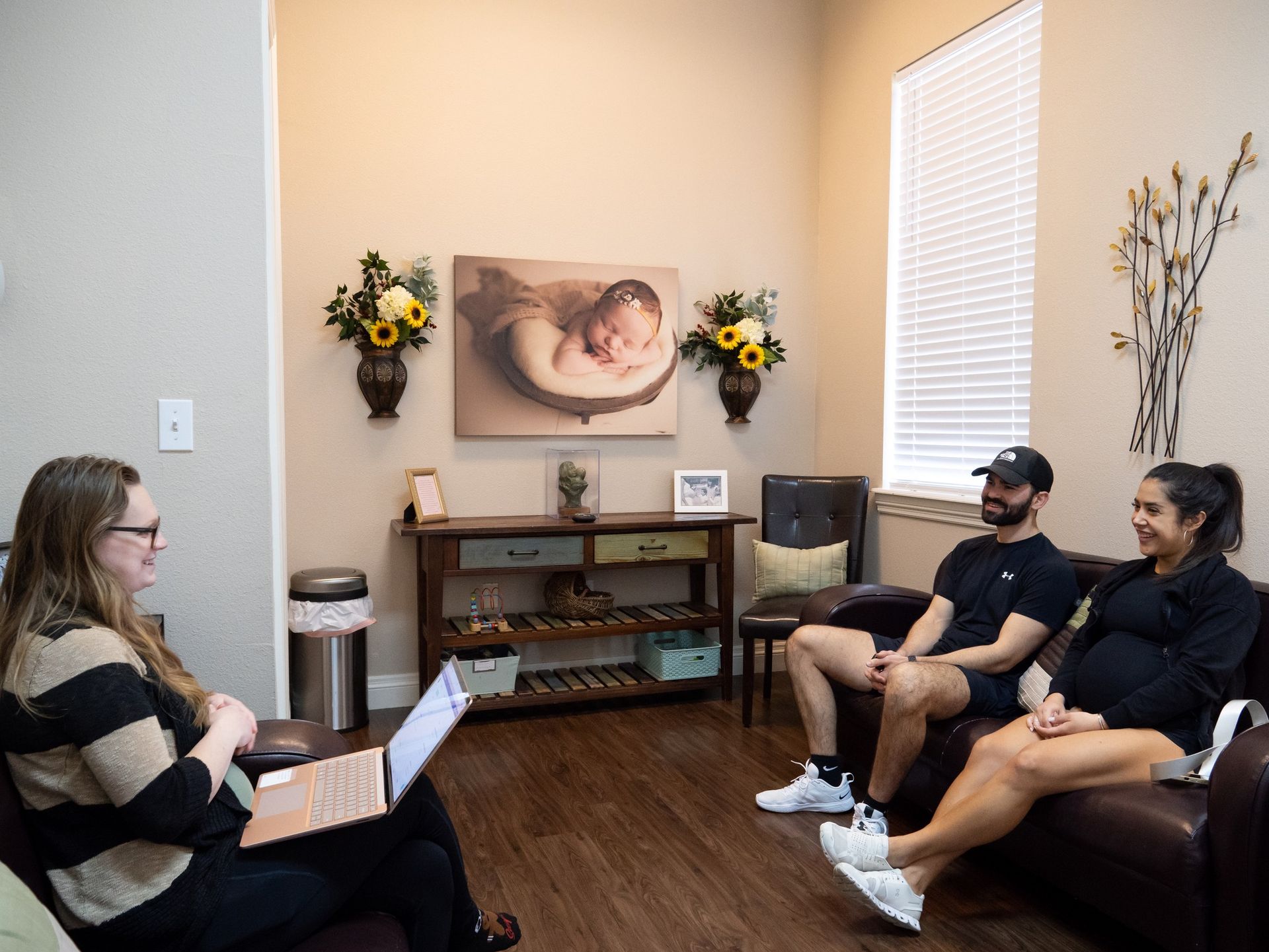 A woman in a striped sweater holds a laptop across from a couple seated on a couch in a professionally decorated office.