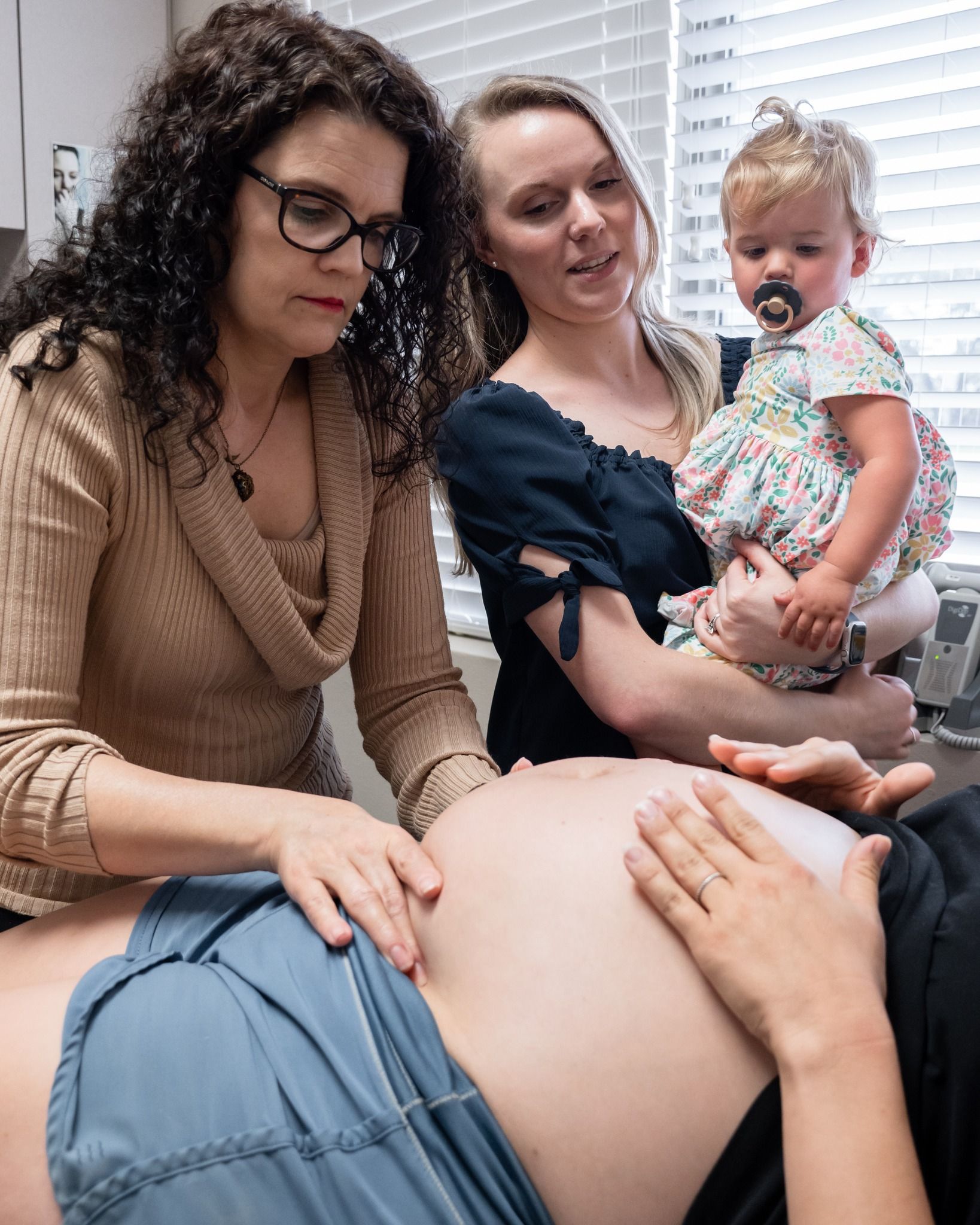 A healthcare provider examines a pregnant person’s abdomen while a person holds a child nearby.