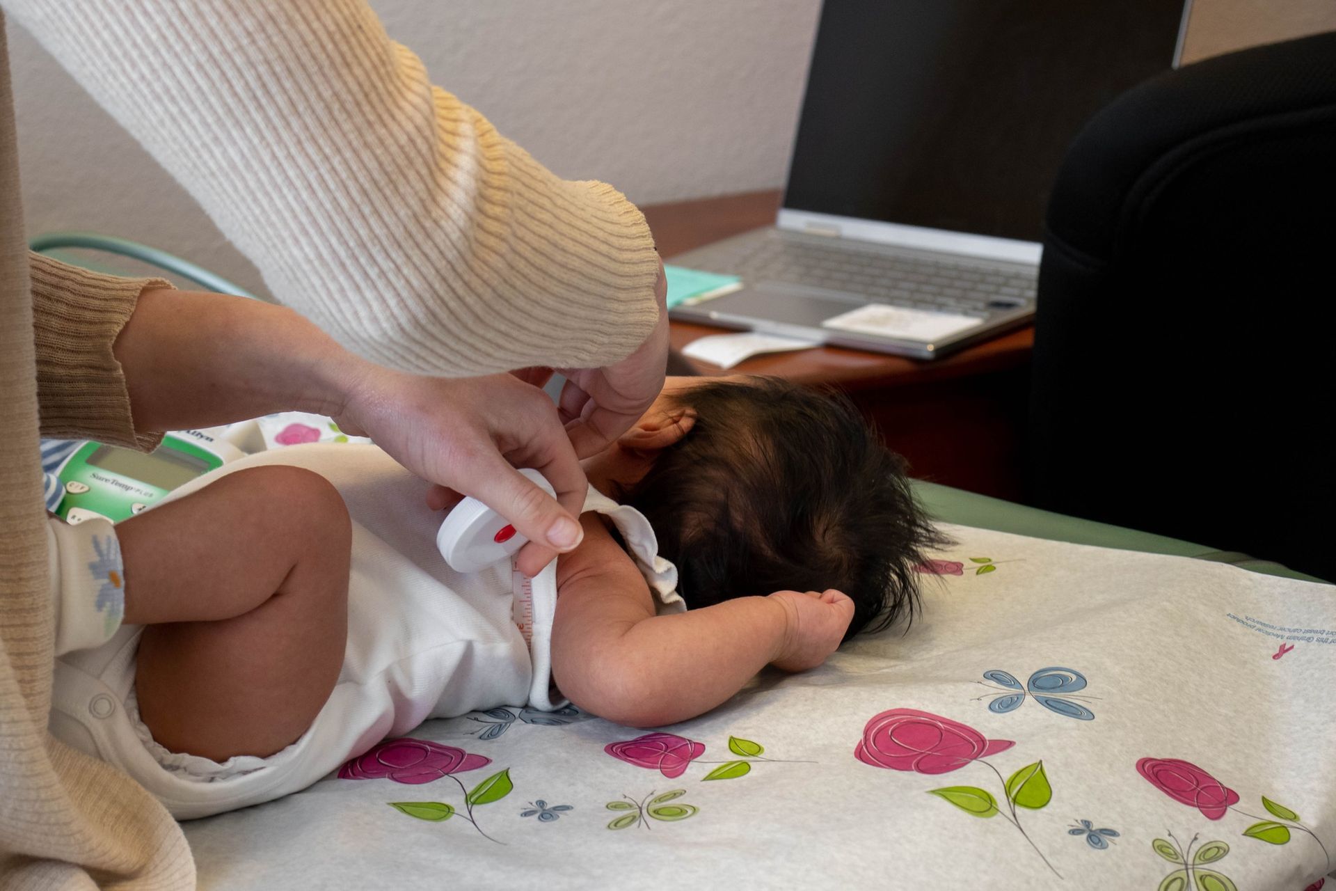 An adult uses a stethoscope to listen to an infant lying on a floral examination table near a laptop.
