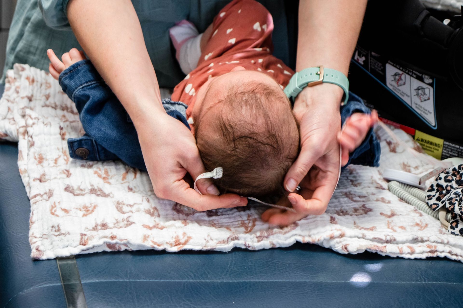 A caregiver adjusts a small sensor on a baby's head while the infant lies on a patterned blanket.