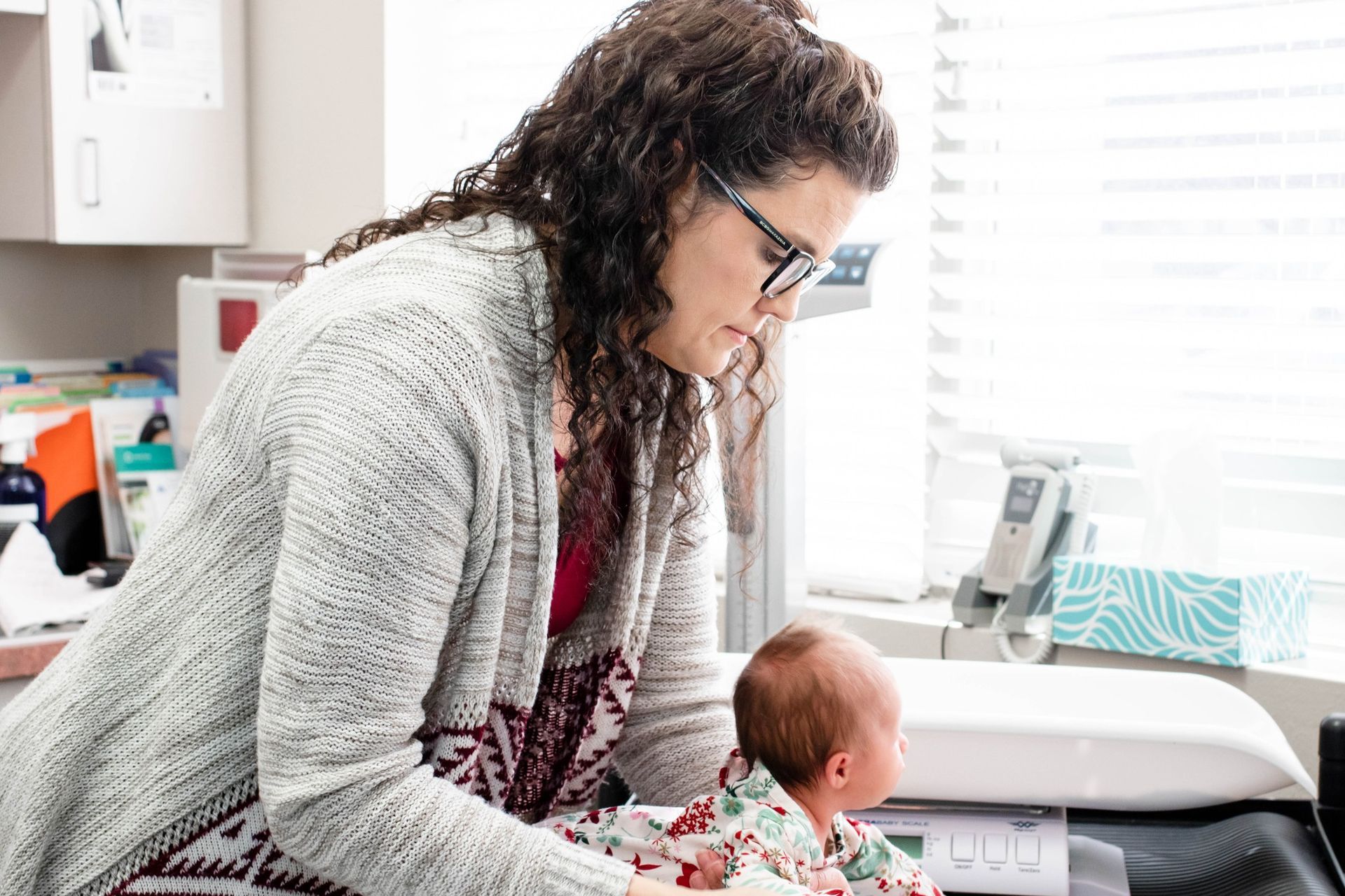 A person with curly dark hair and glasses leans over a baby lying on a medical exam scale in a clinical office.