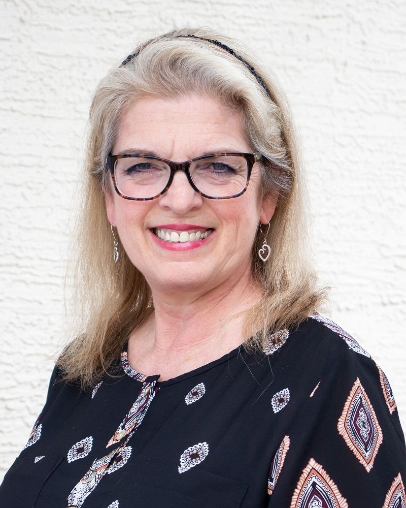 A smiling person with blonde hair, a black headband, and glasses, wearing a black patterned top against a white background.