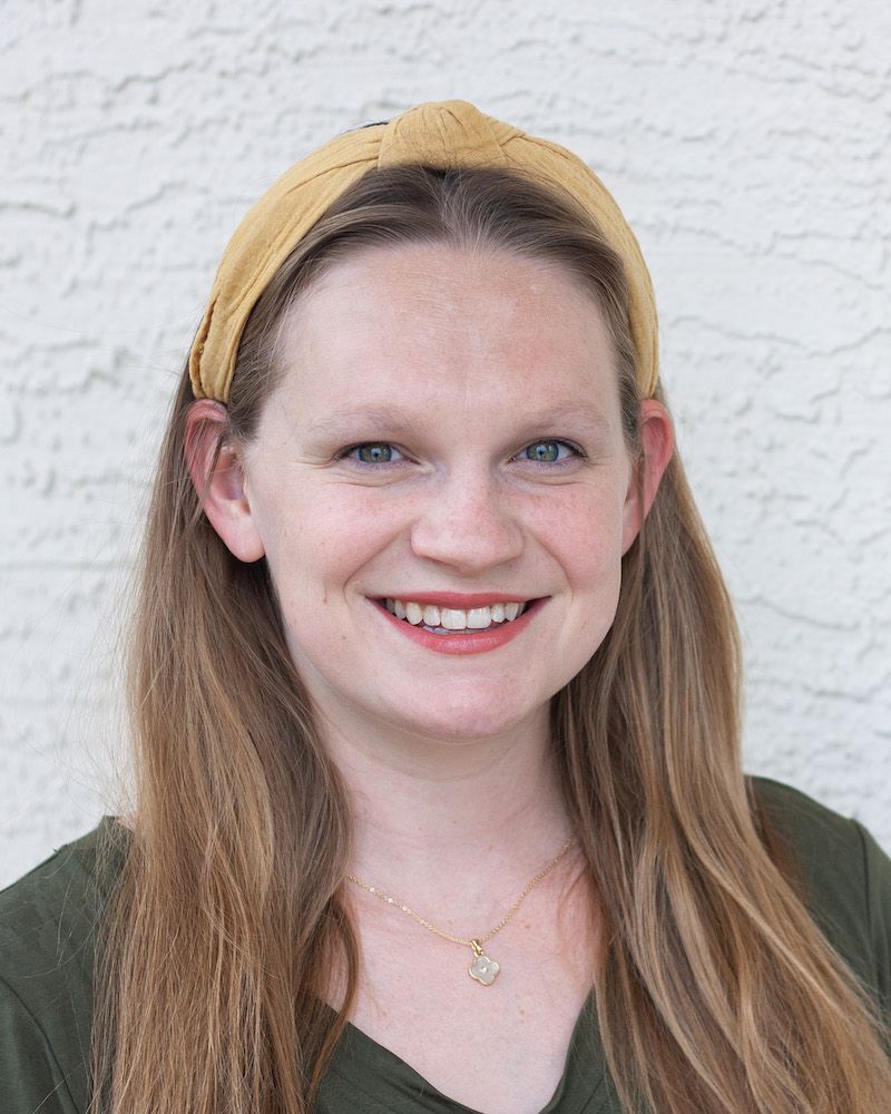 A smiling person with long hair and a mustard-yellow headband wears a green top against a white textured wall.