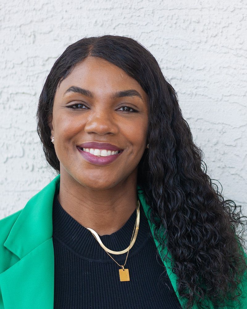 A smiling person with long dark hair, wearing a green blazer and a black shirt with a layered gold necklace.