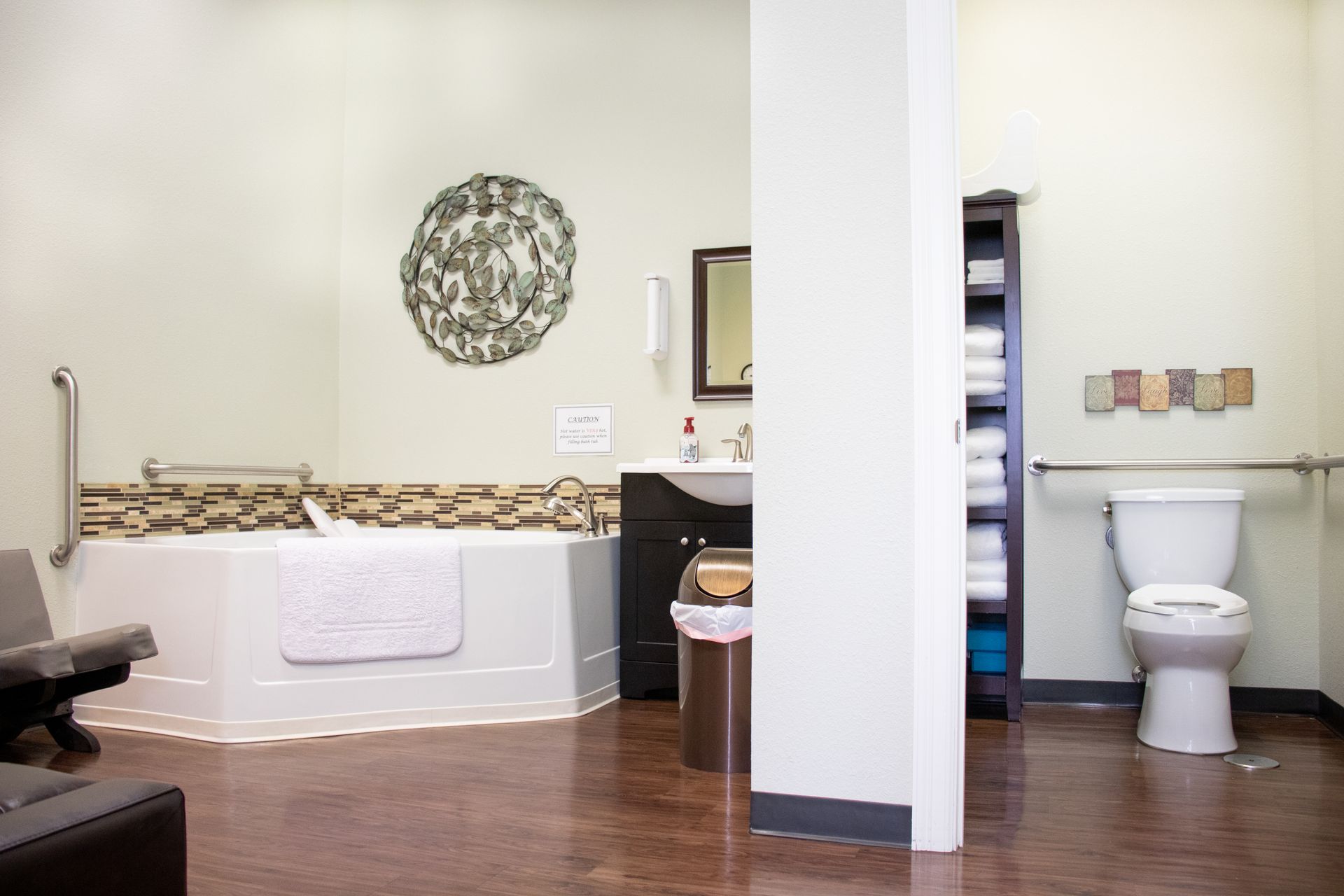 An accessible bathroom featuring a large jetted tub, a sink, a storage tower filled with towels, and a grab-bar toilet.