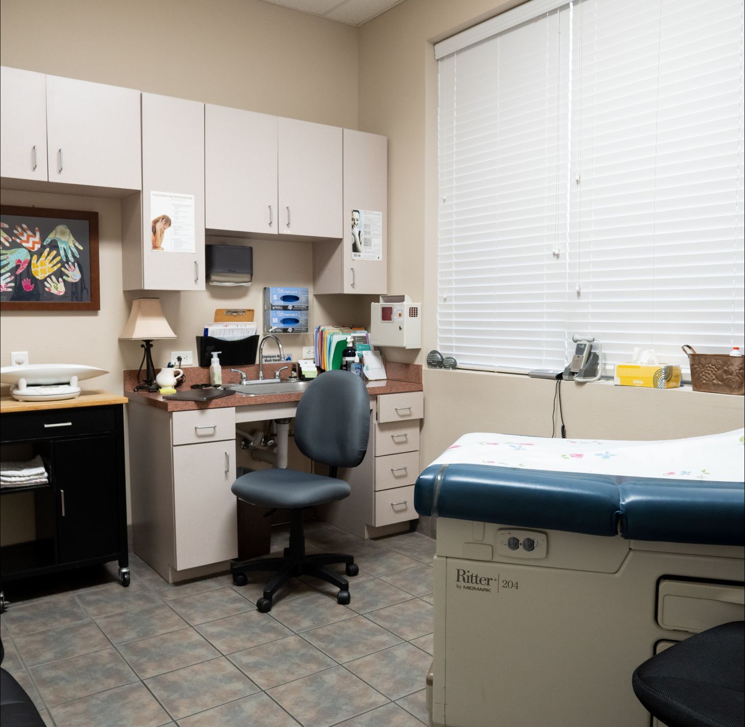 A medical exam room featuring a paper-covered exam table, a gray office chair, cabinetry, and a framed handprint artwork.