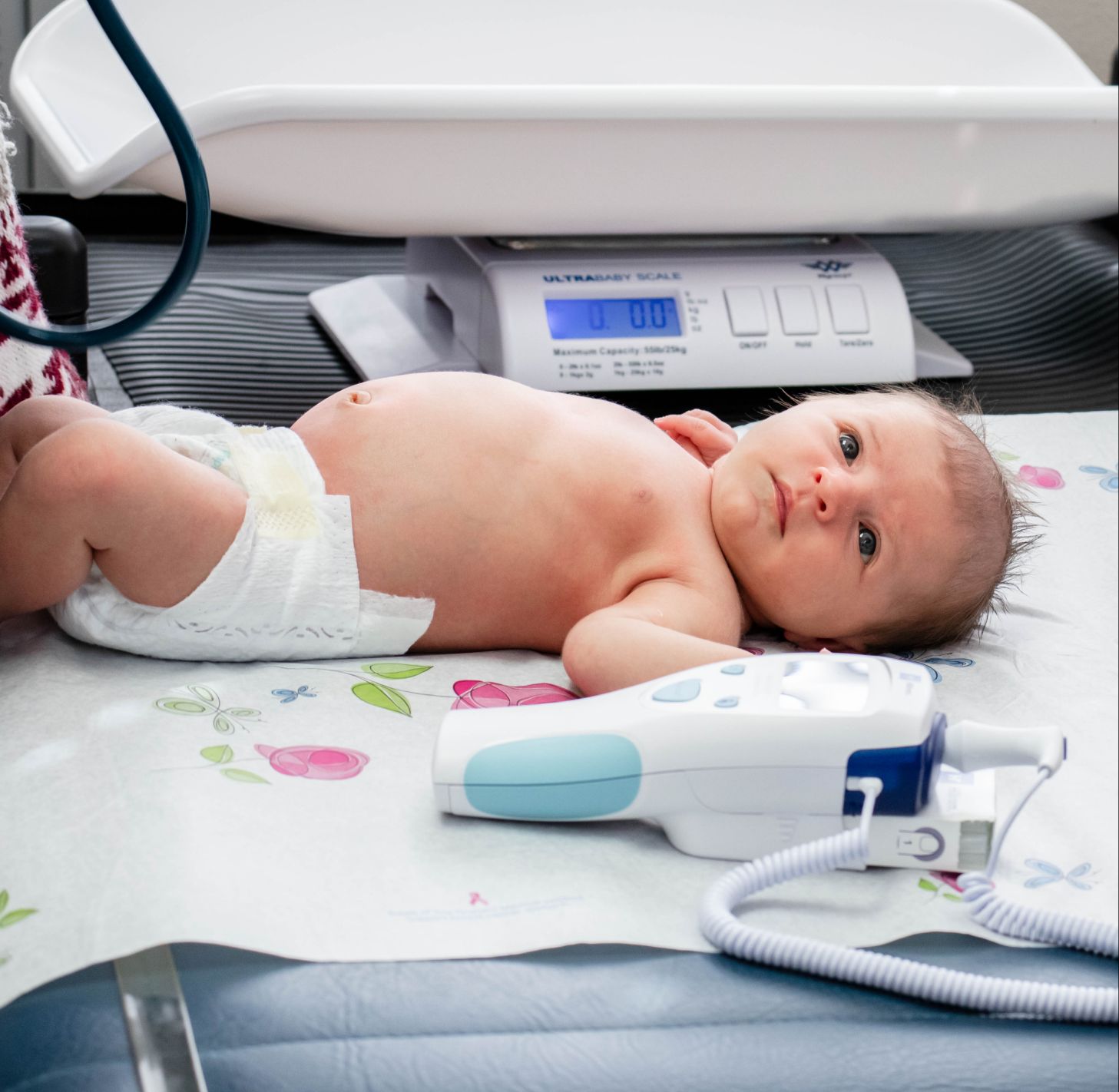 A newborn lies on an exam table next to medical equipment, including a scale and a blood pressure monitor.