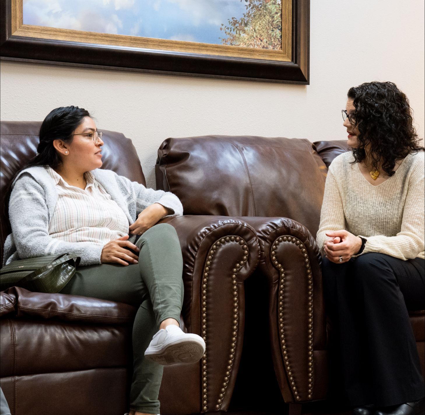 Two people sit in brown leather armchairs, facing each other and talking in a room with a framed painting on the wall.