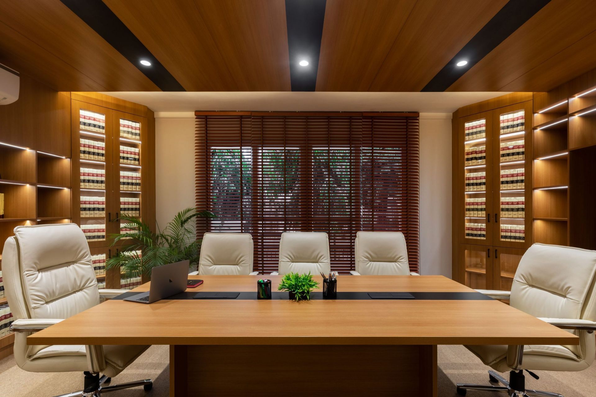 A wood-paneled office featuring a long conference table with five white leather chairs, set against shelves and a window.