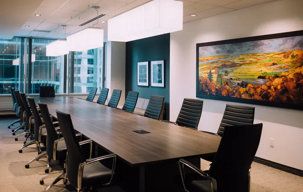 A modern boardroom featuring a long wooden conference table surrounded by black chairs under bright, rectangular lights.