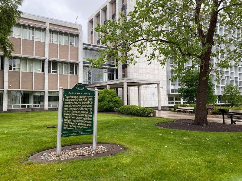 A green historical marker stands on a grassy lawn in front of a modern, multi-story office building.