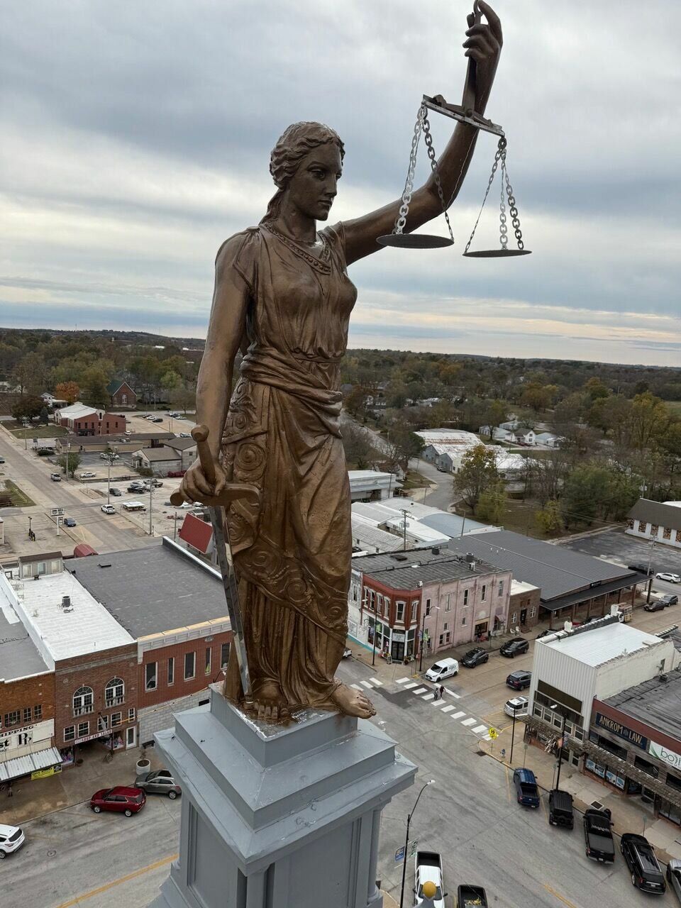 Bronze statue of Lady Justice holding scales and a sword atop a courthouse pedestal, overlooking a town street.