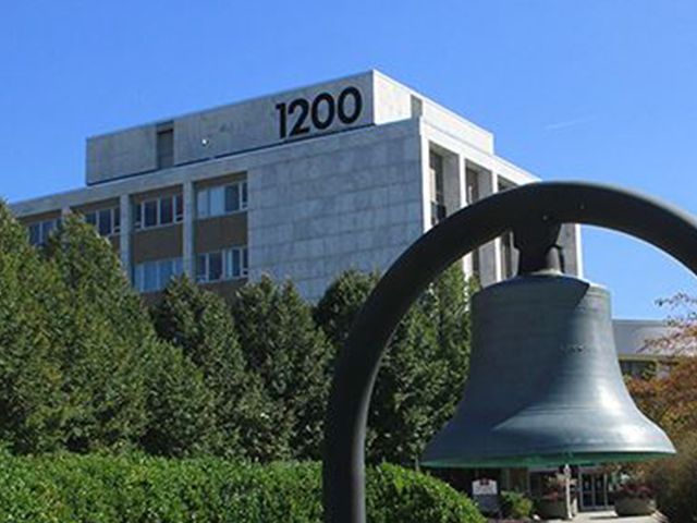A metal bell hangs in the foreground, with a large, light-colored building labeled 
