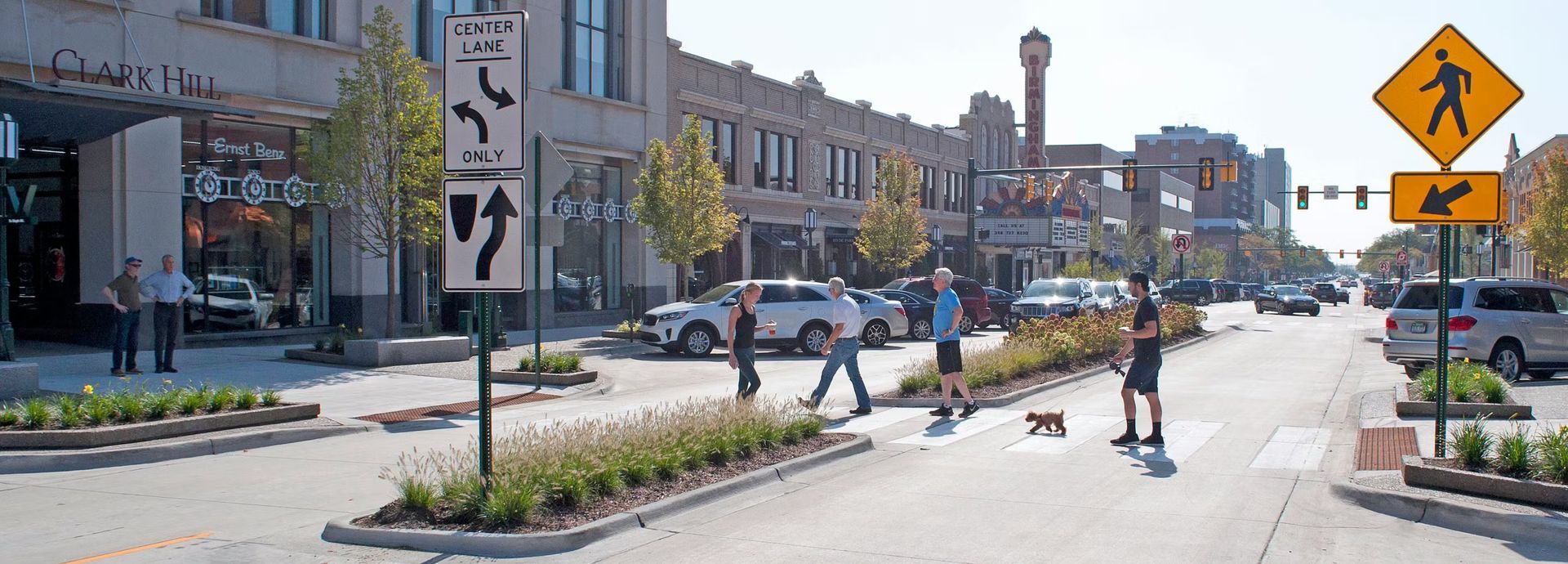 Pedestrians cross a downtown street with a central planted median, traffic signs, and businesses in the background.