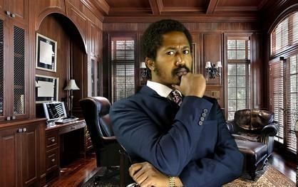 A person in a dark suit with their hand on their chin stands in a wood-paneled office.