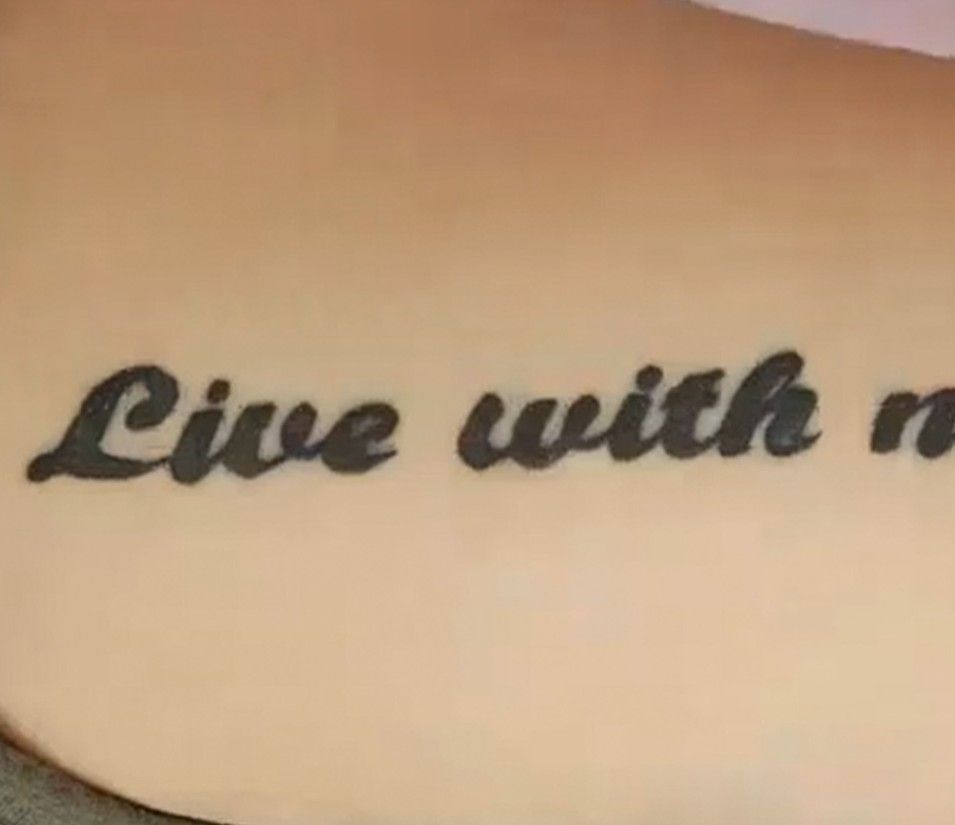 Black script tattoo reading “Live with n” on tan skin.