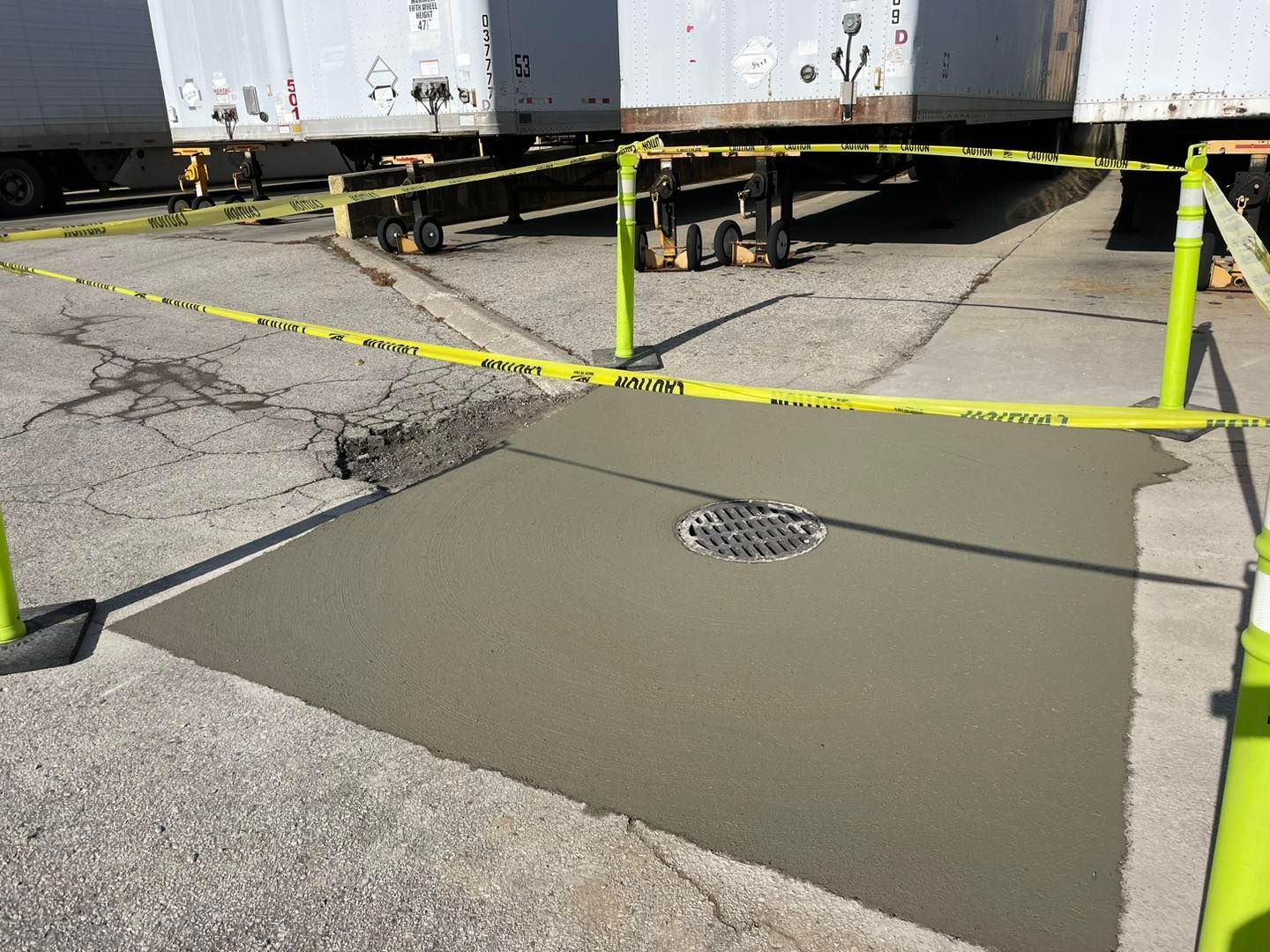 Newly poured concrete patch with drain, cordoned off with yellow caution tape and posts near semi-truck trailers.
