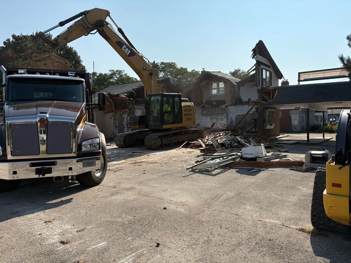 An excavator demolishes a building, loading debris into a truck, under a sunny sky.