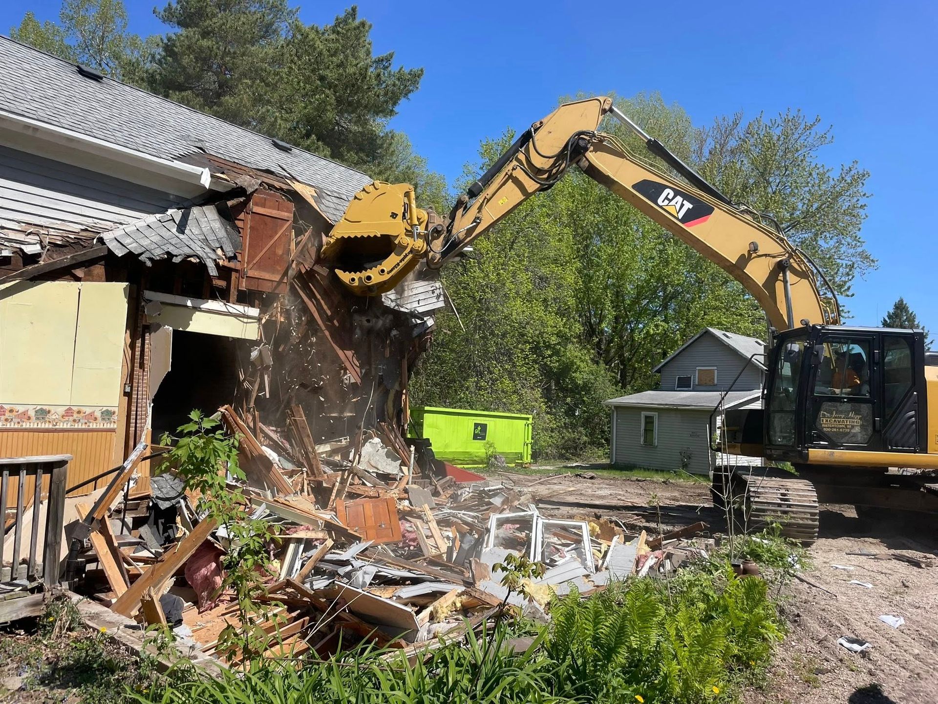 Excavator demolishing a house, tearing off the roof and walls; debris scattered in a yard.