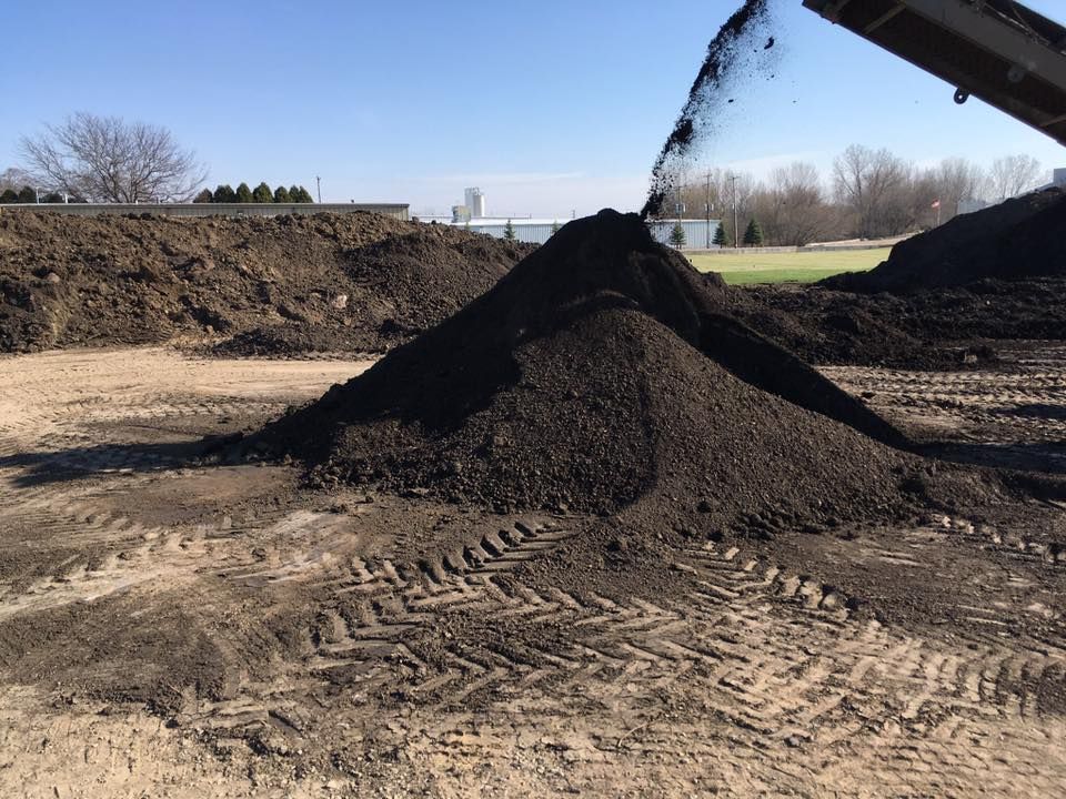 Dark soil pouring from a machine, forming a large pile on a dirt surface.