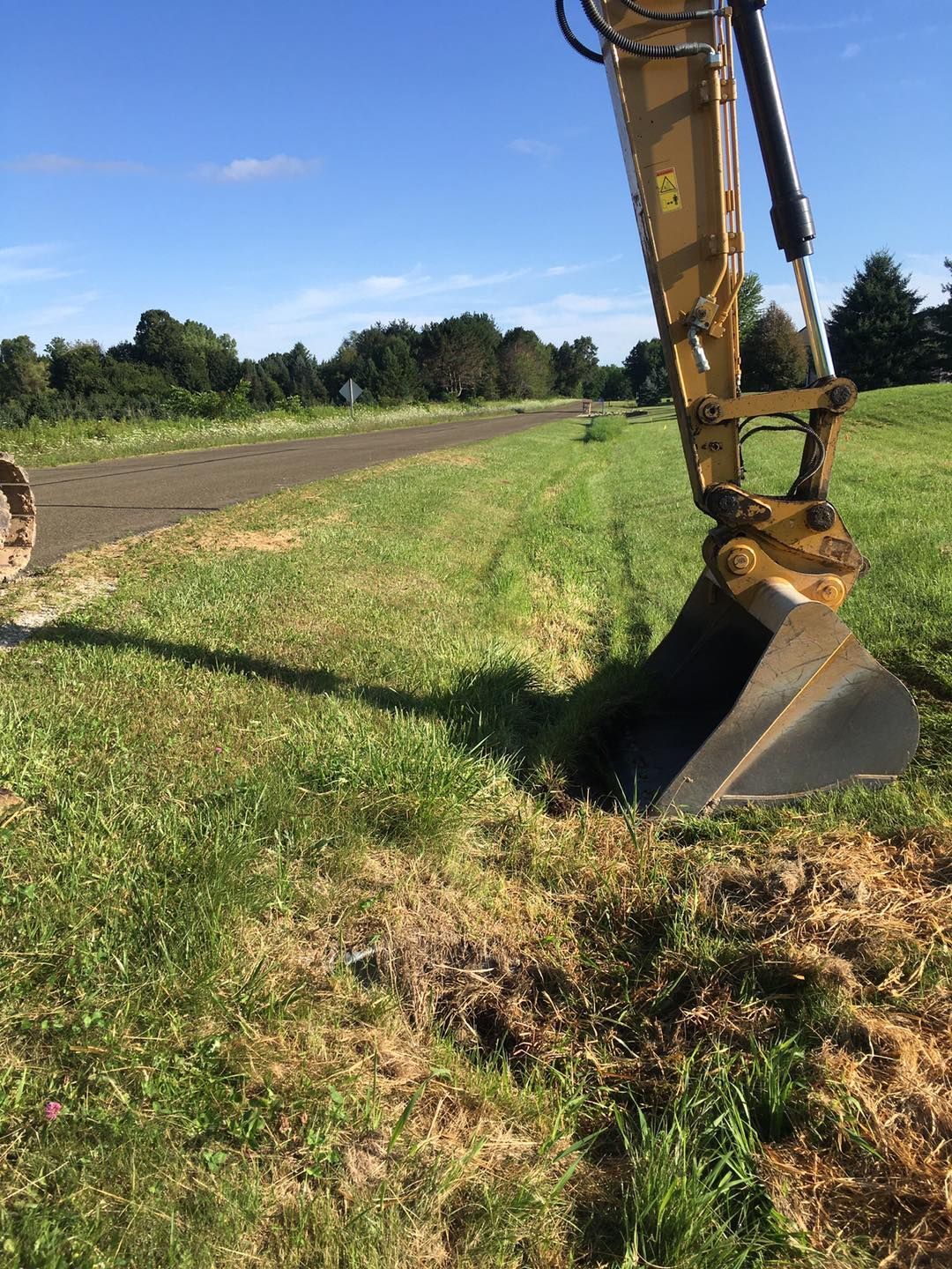 An excavator digging a trench in a grassy field next to a path, under a blue sky.