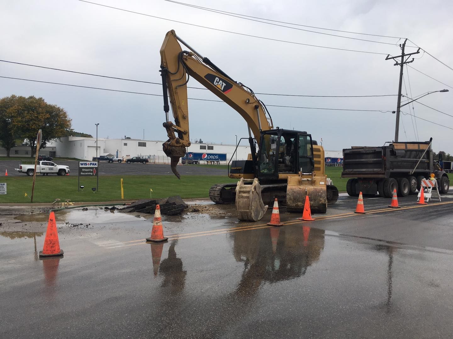 Excavator digging in wet asphalt, orange cones, dump truck, power lines, cloudy day.