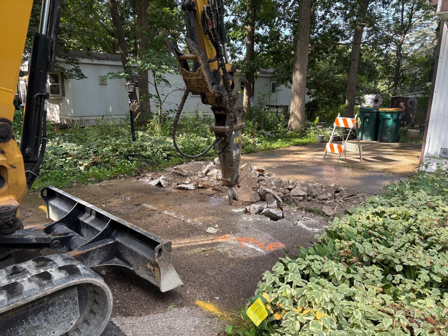 An excavator breaking up concrete in a residential area. Trees, debris, and a barrier visible.