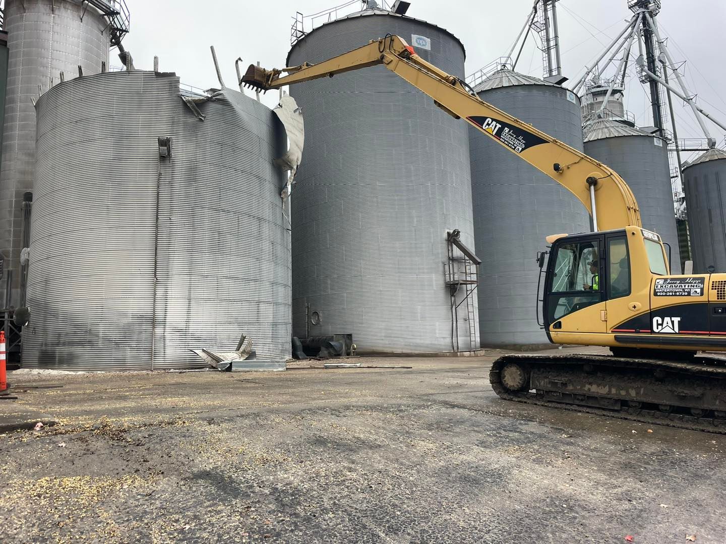 Excavator demolishing grain silo; the silo shows a rippled pattern of damage.