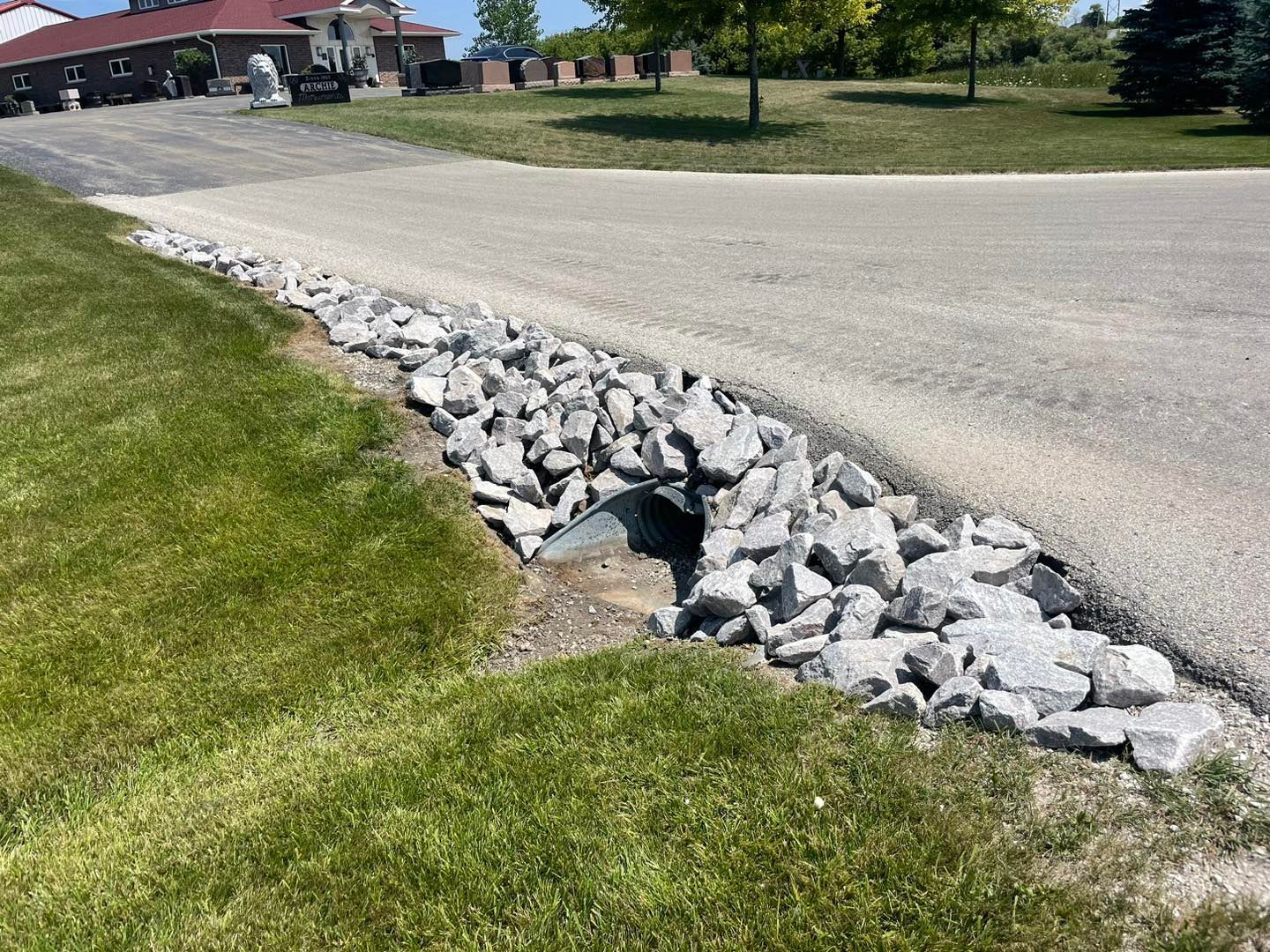 Gravel-lined ditch alongside asphalt road, leading to a culvert under the road, next to a grassy area.
