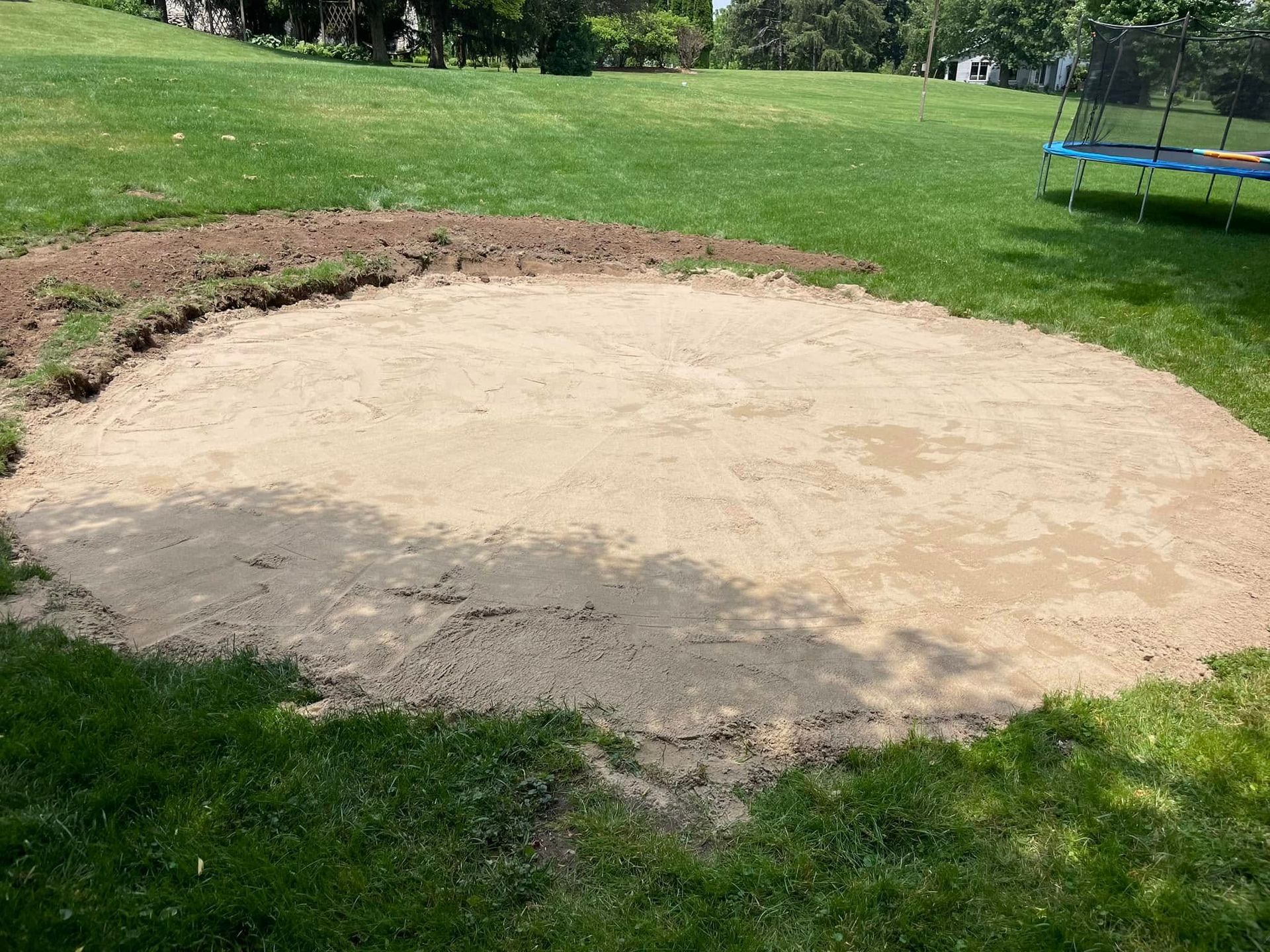 Round dirt pit, sandy-colored, surrounded by grass. Partially excavated with fresh dirt.