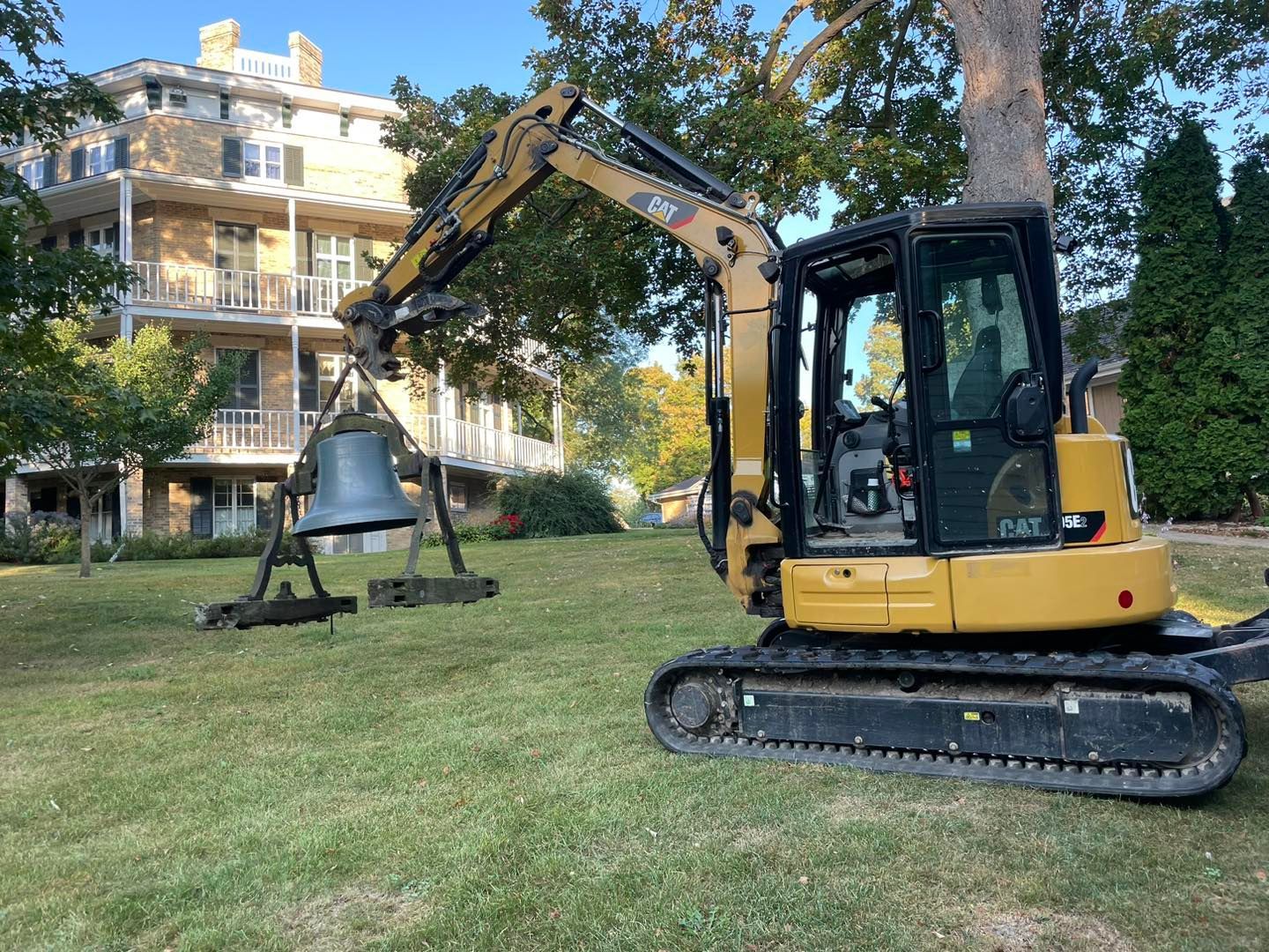 Yellow excavator lifting a large bronze bell on a grassy lawn; building in background.