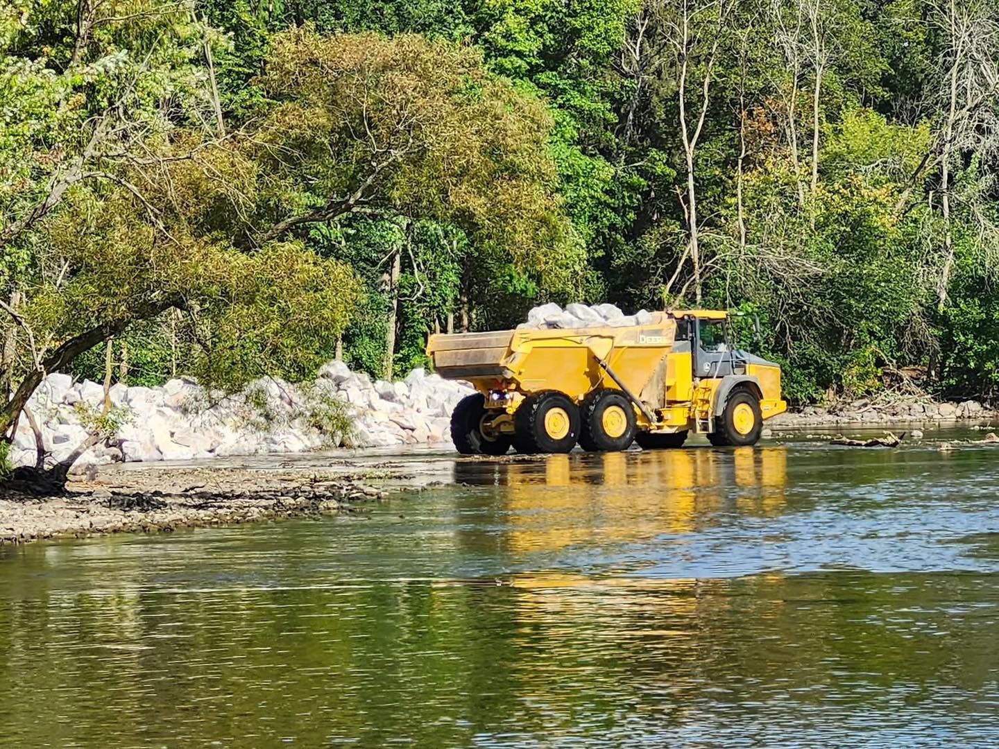 Yellow dump truck driving through shallow water, carrying a load, with trees in the background.