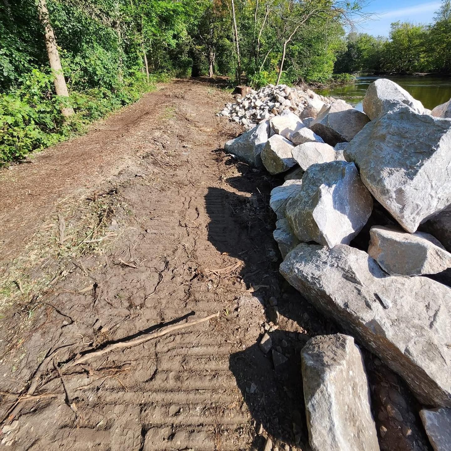 A dirt path next to a river bank reinforced with large rocks and trees.