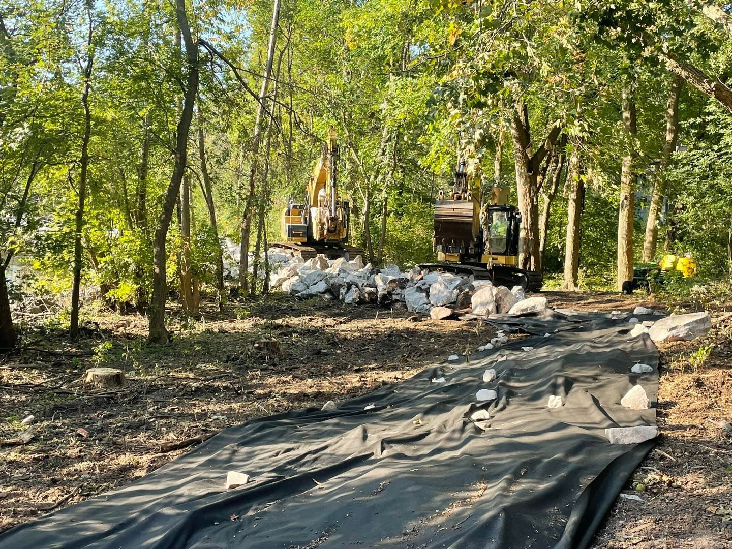 Two excavators on a construction site next to a stream, with a black fabric ground cover and rocks.