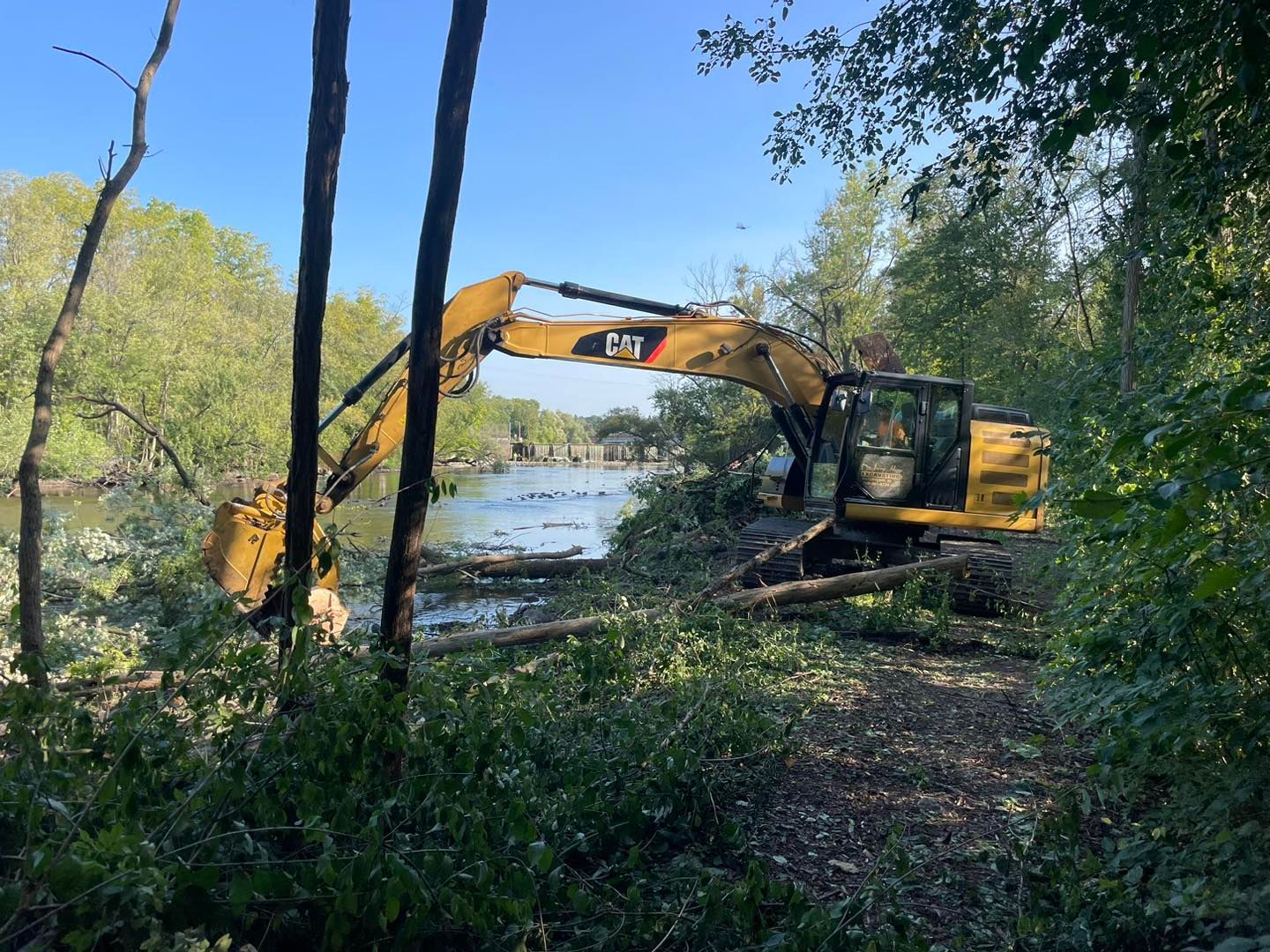Yellow excavator clearing brush near a body of water under a blue sky.