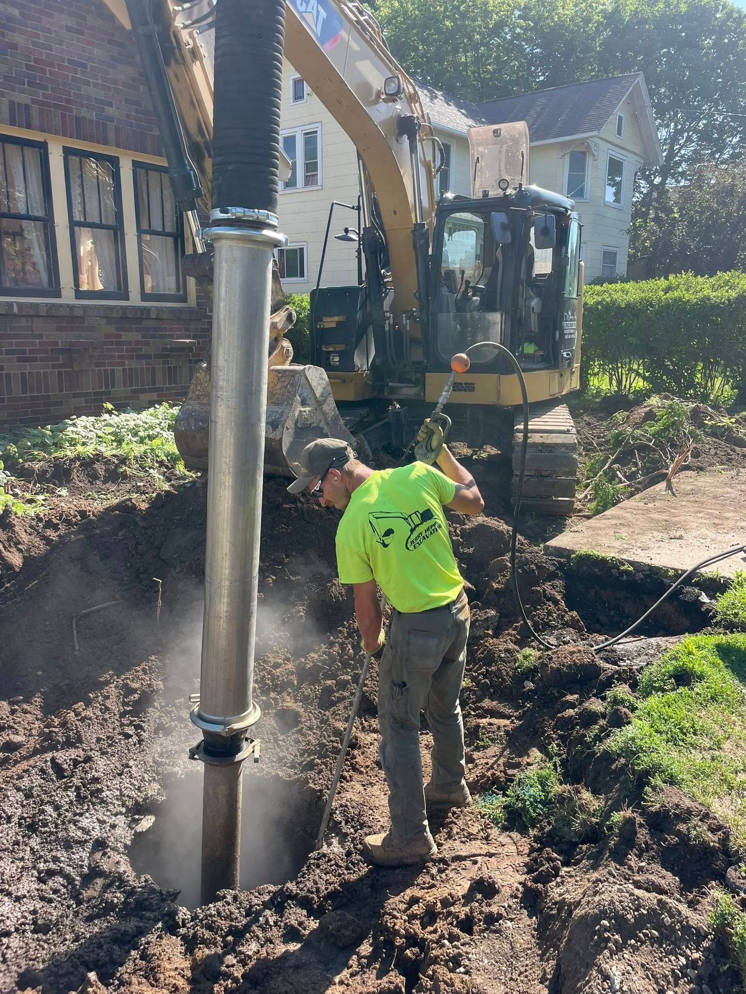 Man in neon green shirt digging near a construction machine; dirt and building in background.