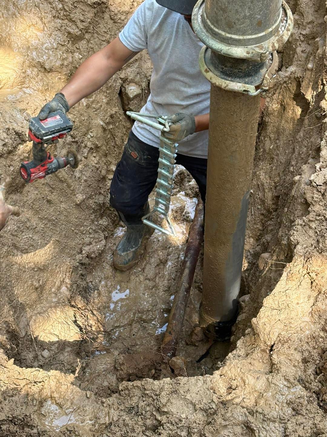 Person in a trench cuts a corroded pipe with a power tool; a metal clamp supports the pipe.