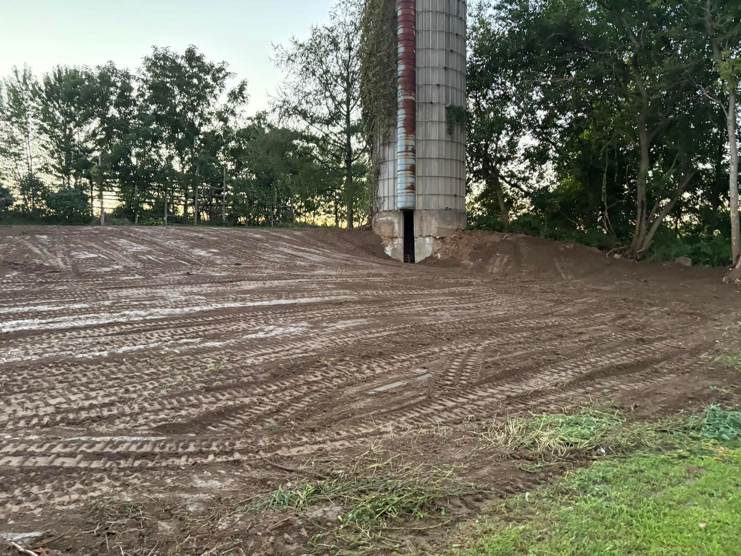 Dirt field with tractor tracks in front of a silo on a farm.