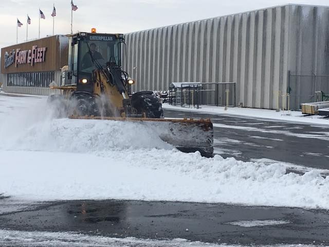 Yellow Caterpillar snowplow clearing snow from a paved lot in front of a building with a large sign.