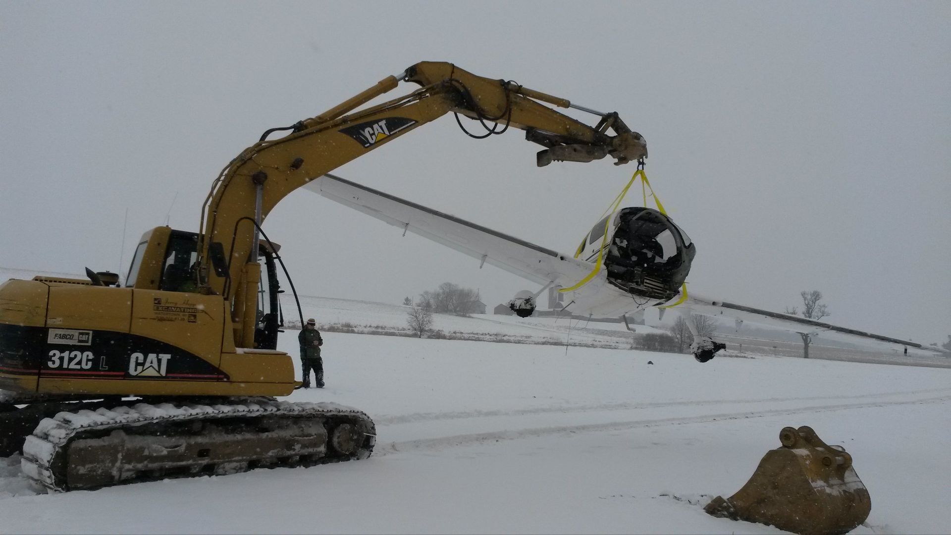 Excavator lifting a crashed, white airplane from a snowy field; a person stands nearby.