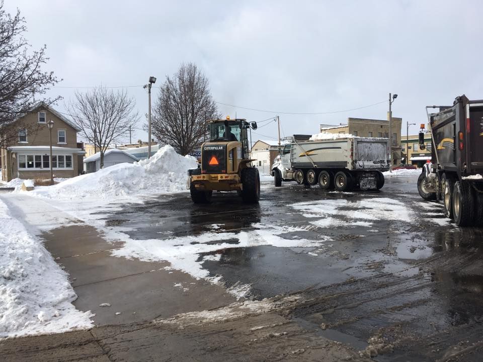 A yellow front-end loader pushing snow into trucks on a wet, snow-covered lot. Buildings in the background.