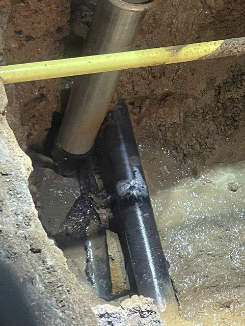 Pipes in a trench, one broken with a black fitting. Water and mud are visible.
