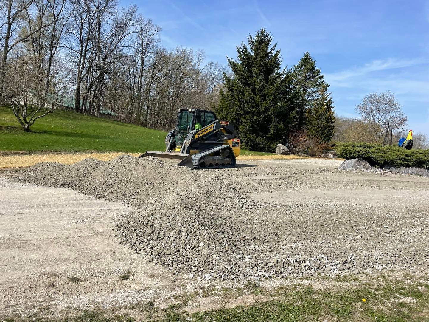 A small, yellow skid steer spreading gravel on a paved area, with green grass and trees in the background.