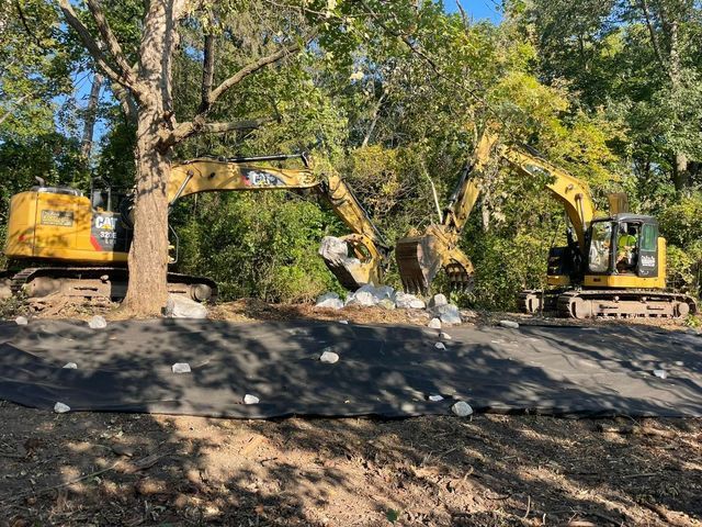 Two yellow excavators on a black surface, placing rocks amidst trees.