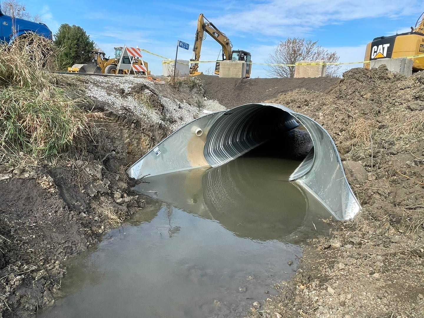 Metal culvert opening into muddy water, construction site with heavy machinery visible.