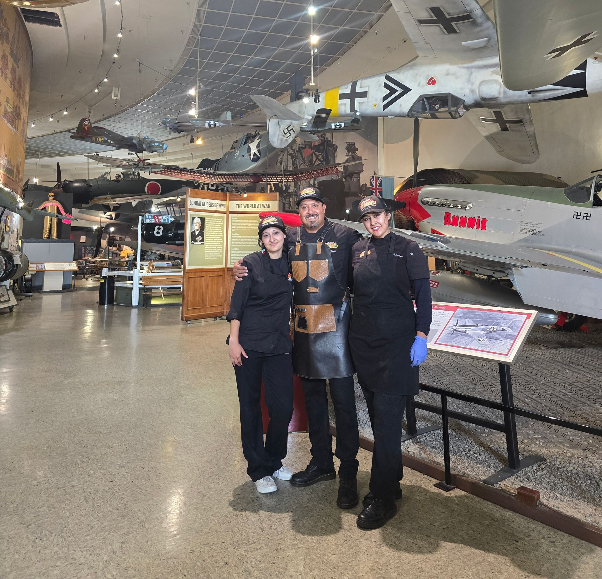 Three people in black uniforms and aprons pose in an aircraft museum.